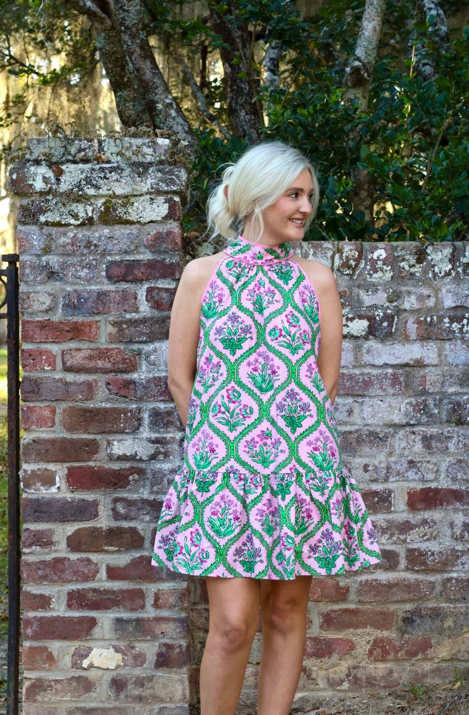 Woman wearing a patterned dress standing against a brick wall with greenery.