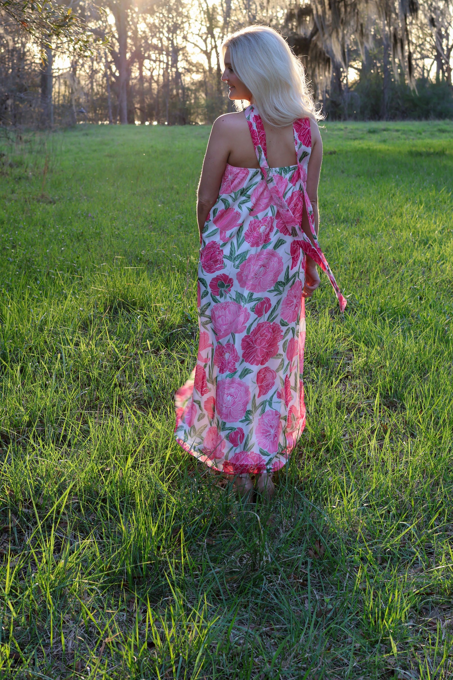 Woman in a pink floral dress standing in a grassy field with trees in the background