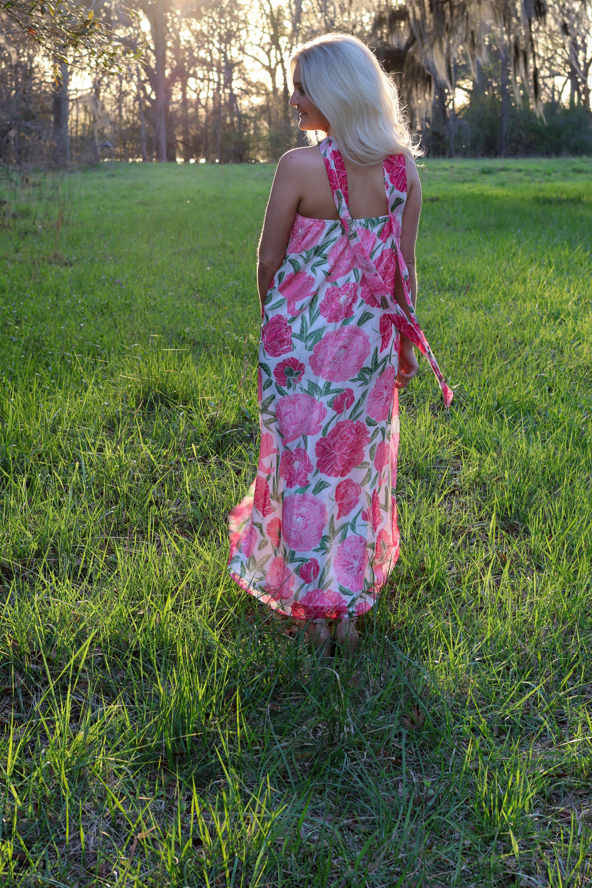 Woman in a pink floral dress standing in a grassy field with trees in the background